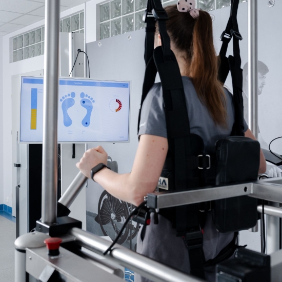 Pacjentka w uprzęży podtrzymującej podczas ćwiczeń w systemie G-EO, z widocznym monitorem prezentującym informacje o ustawieniu stóp.