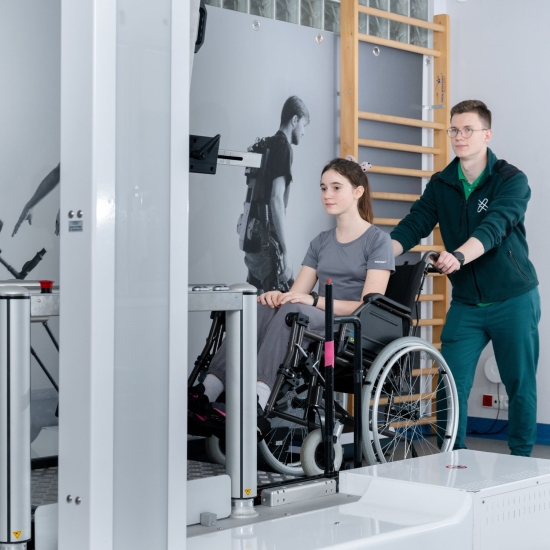 Pacjentka na wózku inwalidzkim podczas przygotowania do ćwiczeń przy stanowisku systemu G-EO z asystą terapeuty.