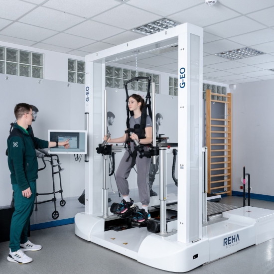 Pacjentka podczas terapii chodu w systemie G-EO pod nadzorem terapeuty, z widocznym panelem sterowania i uprzężą.