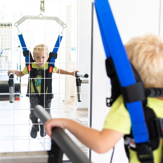 Chłopiec w uprzęży OSTIUM ćwiczący chód przy poręczach rehabilitacyjnych.